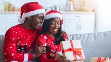 Bir çift, Noel süveteri ve kırmızı Noel Baba şapkaları giyer ve rahat bir koltukta hediyelerini açarken gülümser ve kucaklaşırlar. Festival mevsiminin sıcaklığı odayı dolduruyor..