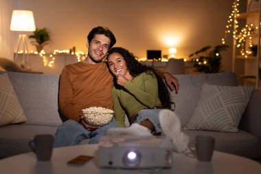 Sıcak bir akşamın tadını çıkarırken, mutlu bir çift kanepede dinleniyor, projektörle film izliyor. Patlamış mısırları paylaşıyorlar, sıcak bir atmosfer yaratıyorlar evde bir hafta sonu için mükemmel..