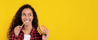 Kıvırcık saçlı genç bir kadın elinde hamburger ve patates kızartmasıyla gülümsüyor. Parlak sarı bir zemin üzerine kurulmuş yemeği için heyecanlı görünüyor..