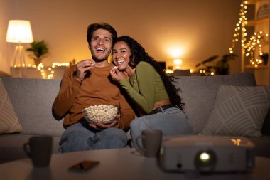 A married couple sits on their sofa indoors, laughing and sharing popcorn while enjoying a comedy film. They are spending a cozy evening together and having a great time.