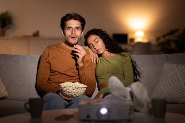 Husband watches a movie using a projector in their living room at night. His wife sleeps beside him on the sofa, showing their different tastes in films during weekend leisure.