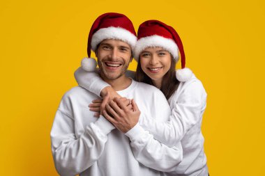 Neşeli bir çift, parlak sarı arka planda kırmızı Noel Baba şapkaları giyerken birbirlerini kucaklıyor. Sıcak bir şekilde gülümserler, tatil sezonunun neşesini somutlaştırırlar, rahat beyaz kıyafetler giyerek..