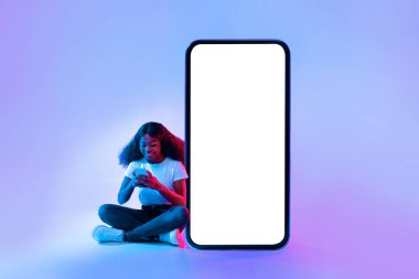 Yerde oturan bir kadın, akıllı telefonuna odaklanmış. Arka planda canlı mavi ve mor ışıklandırma modern ve dijital atmosferi geliştirmektedir..