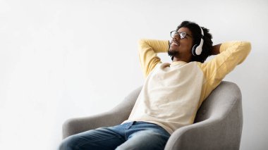 Genç siyah bir adam evdeki bir sandalyede kablosuz kulaklık takarken rahatça dinleniyor. Huzurlu bir hafta sonu boyunca en sevdiği müziği dinlerken gülümsüyor..