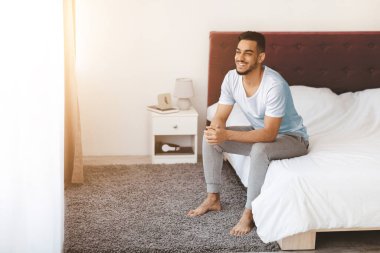 Genç bir adam yatağının kenarında oturuyor, sabah ışığı pencereden içeri girerken gülümsüyor. Rahat bir atmosferi olan, rahat bir yatak odasında rahat rahat giyiniyor..