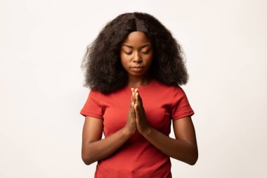 Sakin bir Afrikalı Amerikalı kadın parlak bir stüdyoda elleri sımsıkı bağlı ve gözleri kapalı duruyor. Odaklanmış ve huzurlu görünüyor. Bu huzurlu ortamda bir an için dua ve yansıma yaşıyor..