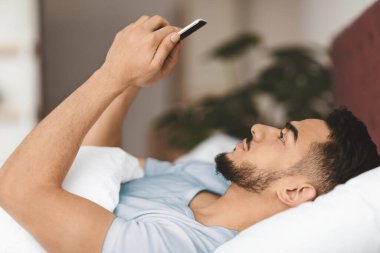 A young man with a beard relaxes in bed while checking his smartphone. Soft morning light filters into the room, creating a calm and cozy atmosphere as he scrolls through his device.