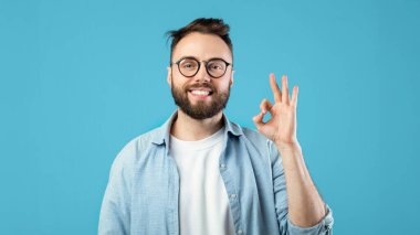 A man with glasses and a beard smiles while making an ok sign with his right hand. He wears a light shirt and stands in front of a vibrant blue backdrop, conveying positivity.