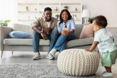 Küçük siyah çocuk oturma odasında yürüyor, garip bir şekilde bir puf 'a dokunuyor. Mutlu Afrika kökenli Amerikalı ebeveynler çocuklarının oyun oynamasını izleyip, rahat bir ev ortamında bağımsızlığı teşvik ediyorlar..