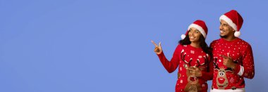 Noel süveteri ve Noel Baba şapkası giyen neşeli bir çift birlikte poz verirken coşkulu görünüyorlar. Noel ruhunu kutluyorlar, parlak mavi bir arka plana karşı..