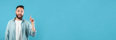 Sakallı ve kısa saçlı neşeli bir adam parlak mavi bir arka planda duruyor. Büyük bir gülümsemeyle yukarıyı işaret ediyor. Hevesli ya da yeni bir fikir öneriyor..