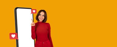 Kırmızı elbiseli gülümseyen bir kadın boş ekrana işaret eden büyük bir akıllı telefonun yanında duruyor. Kalpli sosyal medya simgeleri ortaya çıkıyor ve çevrimiçi müdahale veya etkileşim öneriyor.