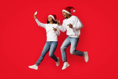 Noel Baba şapkalarını takıp neşeli bir şekilde havaya atlayan bir çift, canlı kırmızı bir zemine karşı, bayram sezonu için mutluluk ve heyecan gösteriyorlar..