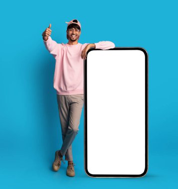 Pembe kazak giyen ve başparmağıyla gülümseyen genç bir adam. Parlak mavi arkaplanı olan boş bir telefona yaslanıyor. Rahat duruşu heyecanı vurguluyor..