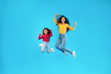 Neşeli bir anne ve kız yüzlerinde gülümsemeyle havaya sıçrıyorlar. Parlak, renkli kıyafetler giyiyorlar ve canlı mavi bir arka plana karşı neşeli bir anın tadını çıkarıyorlar..