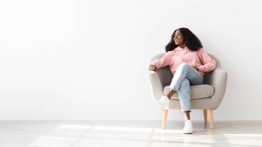 Genç bir kadın, pembe kapüşonlu ve açık renk kotlu bir sandalyede rahatça oturuyor. Neşeli görünüyor, minimalist uzayın parlak, havadar atmosferinin tadını çıkarıyor..