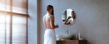 Havluya sarılı şık bir banyoda duran bir adam güne hazırlanıyor. Modern dekorun ve hafif ışığın arasında kendini tımar ederken aynadaki yansımasına bakar..