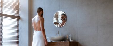 Çağdaş bir banyoda duran, havluya sarılmış ve aynadaki yansımasına bakan bir adam. Sabah bakımıyla meşgul gibi görünüyor..