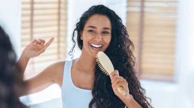 Kıvırcık saçlı neşeli bir kadın yansımasına gülümserken saçını tarıyor. Güneş ışığı ahşap panjurların arasından süzülür ve odada sıcak bir atmosfer yaratır..