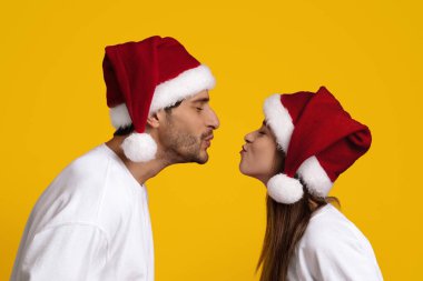 Genç bir çift burunları birbirine değmek üzereyken karşı karşıya duruyor, ikisi de kırmızı Noel Baba şapkaları takıyor..