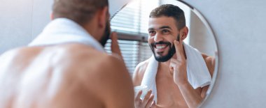 Genç bir adam yuvarlak bir banyo aynasının önünde duruyor, yüzüne bir cilt bakım ürünü sürerken gülümsüyor. Boynuna bir havlu takıyor, özgüvenini gösteriyor..