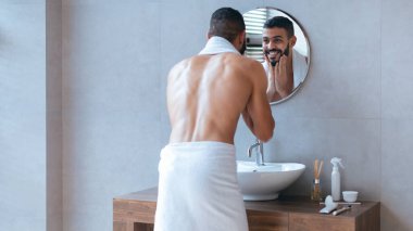 Modern bir banyoda duran bir adam aynadaki yansımasını havluyla kontrol ediyor. Sahne, onun sabah bakım rutinini bir pencereden gelen doğal ışıkla yakalıyor..