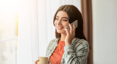 Genç bir kadın pencerede duruyor, telefonda konuşurken gülümsüyor. Bir elinde fincanı tutuyor, odasında sıcak ve aydınlık bir atmosferin tadını çıkarıyor..
