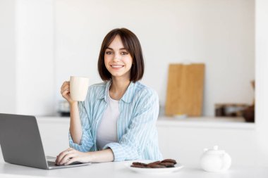 A young woman smiles while holding a cup of coffee, sitting at a table with a laptop. Cookies are nearby, and the kitchen has a fresh, clean design. Natural light fills the space.