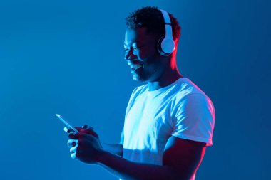 A young man smiles while using his smartphone, listening to music through white headphones. The background features vibrant blue lighting, creating a lively atmosphere.