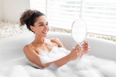 Kabarcık dolu bir küvette oturan bir kadın, kendini aynada incelerken gülümsüyor. Yumuşak ışık pencereden süzülerek sakin bir atmosfer yaratır..