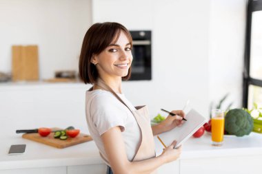 Önlüklü genç bir kadın parlak bir mutfakta duruyor, gülümsüyor ve not defterine yemek tarifi fikirleri yazıyor. Onun yanındaki tezgahta taze sebzeler var..