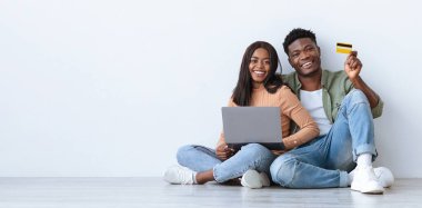 Genç bir çift online alışveriş yaparken yerde oturup gülümsüyor. Ortaklardan biri kredi kartı tutarken diğeri dizüstü bilgisayar kullanarak evde neşeli bir atmosfer yaratıyor..