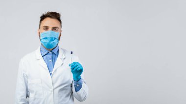 Koruyucu maskeli Millennail erkek doktor ve steril çubuk tutan eldivenler Coronavirus PCR testi için gri stüdyo arka planında. Geniz ve oral covid tanı prosedürü için tüplü genç doktor.