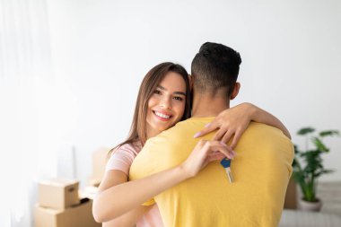 Neşeli bir çift, yeni evlerinde dururken kucaklaşır. Bir ortak, paketleri açık kutular ve kapalı bitkilerle çevrili bir şekilde gülümserken anahtarları tutuyor..