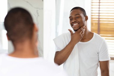 Bir adam aynanın önünde duruyor, omuzunda bir havluyla kendini tımar ederken gülümsüyor. Doğal ışık banyoyu doldurur ve sıcak bir atmosfer yaratır..