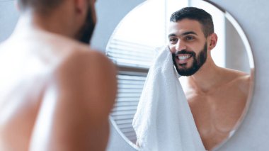 Bir adam banyo aynasının önünde duruyor, yüzünü beyaz bir havluyla kuruturken gülümsüyor. Aydınlık ve temiz ortam, sabah rutininin tımar ve kendine bakmayla ilgili olduğunu gösteriyor..