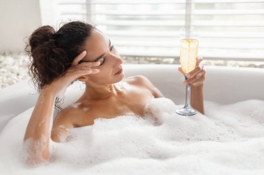 Bir kadın köpüklü bir banyoda bir bardak soda içerken huzurlu bir anın tadını çıkarır. Güneş ışığı pencereden süzülerek sakin atmosferi arttırır..