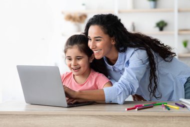 A mother and her daughter sit together at a table, engaged in an enjoyable learning activity on a laptop. They share smiles and laughter, creating a joyful atmosphere.