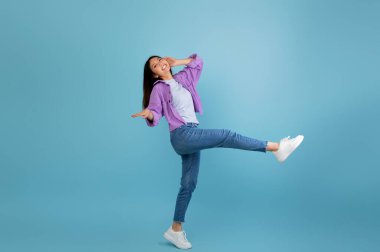 Uzun saçlı genç bir kadın mavi arka planda neşeli bir dans pozu veriyor. Neşeli ve tasasız görünüyor, neşeli havasını ve şık kıyafetini sergiliyor..