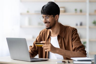 Gözlüklü genç bir adam çalışma alanında dizüstü bilgisayar kullanırken gülümsüyor. İnternetten alışveriş yaparken elinde bir kredi kartı tutuyor ve rahat bir öğleden sonranın tadını çıkarıyor..