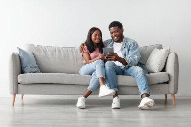Hafta sonu eğlencesi. Evlerinde tablet kullanan, internete giren veya ne alacaklarını seçen, oturma odasındaki rahat koltukta oturan mutlu ve sevecen genç siyah çift. Kadını kucaklayan adam, bedava kopya alanı.