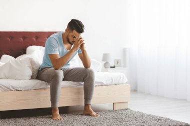 Açık mavi gömlekli bir adam, elleri birbirine kenetlenmiş bir şekilde yatağın kenarına oturur. Derin düşüncelere dalmış, parlak ve havadar bir oda ile çevrili yumuşak doğal ışığın içeri aktığı.