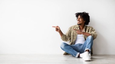 Genç ve neşeli bir Afro-Amerikan erkek yerde oturuyor, açık tişört ve kot pantolon giymiş. Mutlulukla iki parmakla kenarı işaret ediyor, fikirler için boş alan gösteriyor..