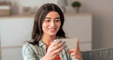 Genç bir kadın modern bir oturma odasında rahat bir şekilde oturuyor, akıllı telefonunu kullanırken gülümsüyor. Güneş ışığı odayı doldurur, sıcak ve davetkar bir atmosfer yaratır..