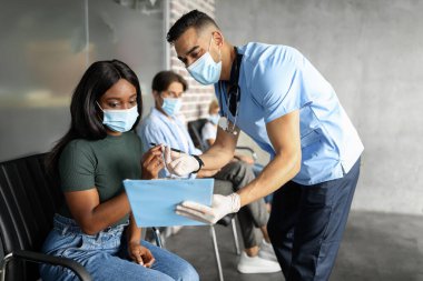 Koruyucu maskeli genç siyahi bayan COVID-19 aşısından önce tıp dosyası imzalıyor ve Orta Doğulu doktorla sohbet ediyor. Aşı noktasında çok ırklı bir grup insan