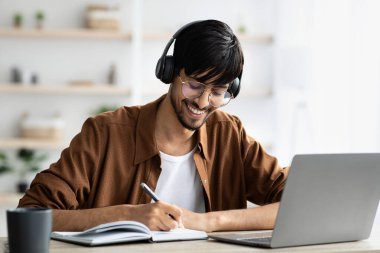 A young man is enjoying his study session at home. He wears headphones, smiles while writing in a notebook, and works on his laptop in a bright, organized space.