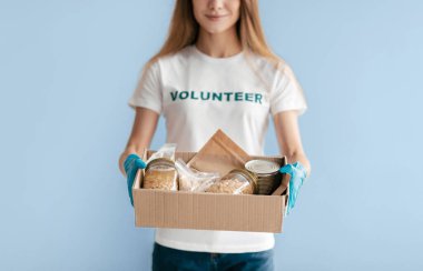 Gönüllü gömleği ve mavi eldivenleri olan uzun saçlı biri açık mavi bir arkaplanda duruyor. Elinde yiyecek dolu bir kutu var, başkalarına yardım etmeye hazır..