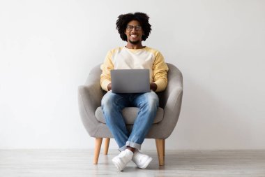 Neşeli genç Afrikalı Amerikalı adam kucağında dizüstü bilgisayarla bir sandalyede rahatça oturuyor. Mesafeli işini seviyor, çalışırken gülümsüyor, rahat bir atmosfer yaratıyor..