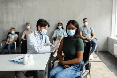 Koruyucu maskeli Afro-Amerikalı genç bayan klinikte COVID-19 'a karşı aşı oluyor. Aşı bekleyen çok ırklı bir grup insan, erkek doktor enjeksiyon yapıyor.