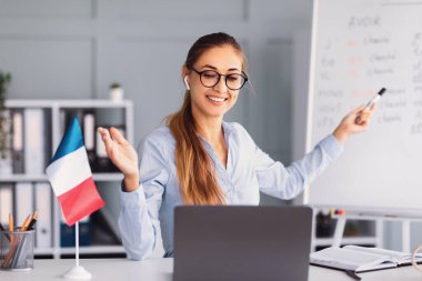 Bir öğretmen gülümsüyor ve internetten bir ders veriyor, beyaz tahta ve Fransız bayrağıyla etkileşimli bir yaklaşım sergiliyor. Samimi çalışma alanı olumlu bir öğrenme ortamı öneriyor..